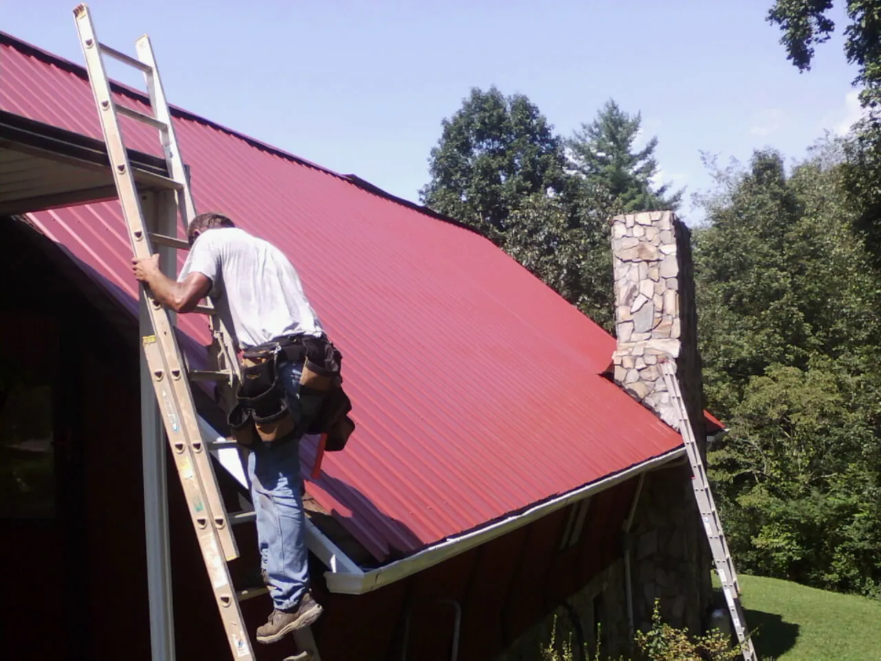Roofer on a ladder beside a red metal roof and stone chimney — Soffit & Fascia Repair by Roofing Brothers in North Yarmouth